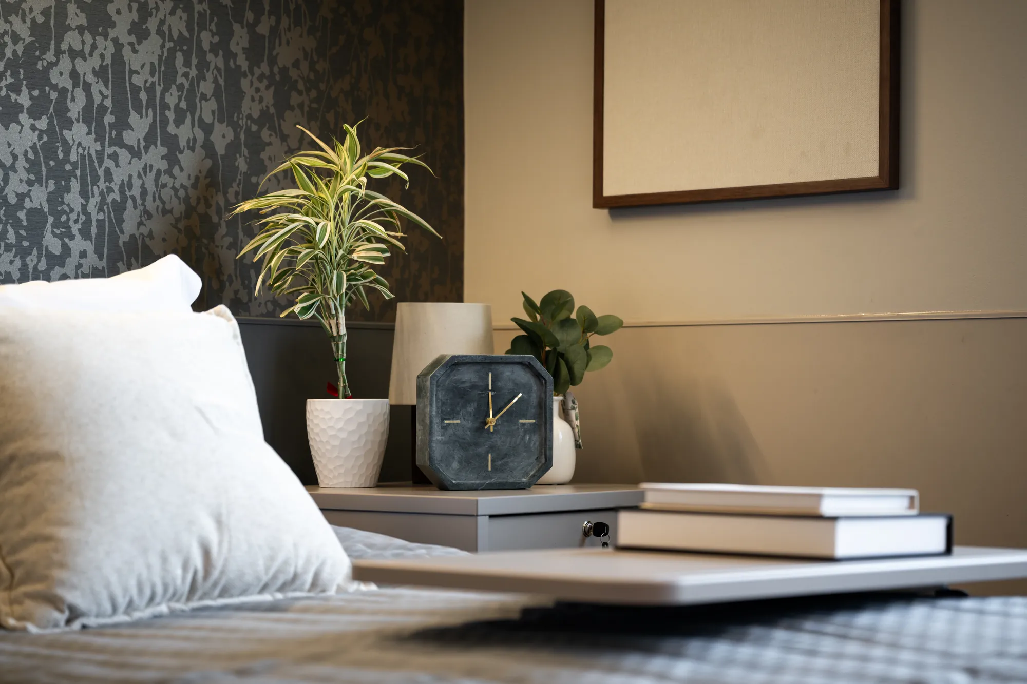 Bedside detail in an Amara resident room: pillows, a clock, a plant and a stack of books