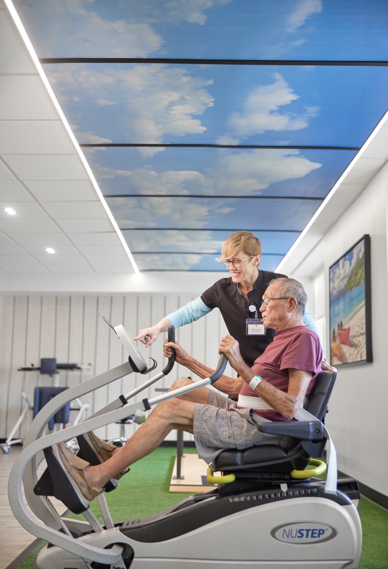 Physical therapist guiding a resident through NuStep cardio under the sky-mural ceiling