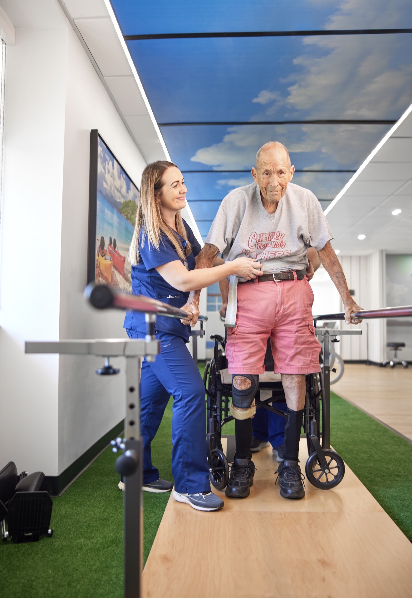 Therapist supporting a resident on parallel bars under the sky-mural ceiling at Village Place rehab gym