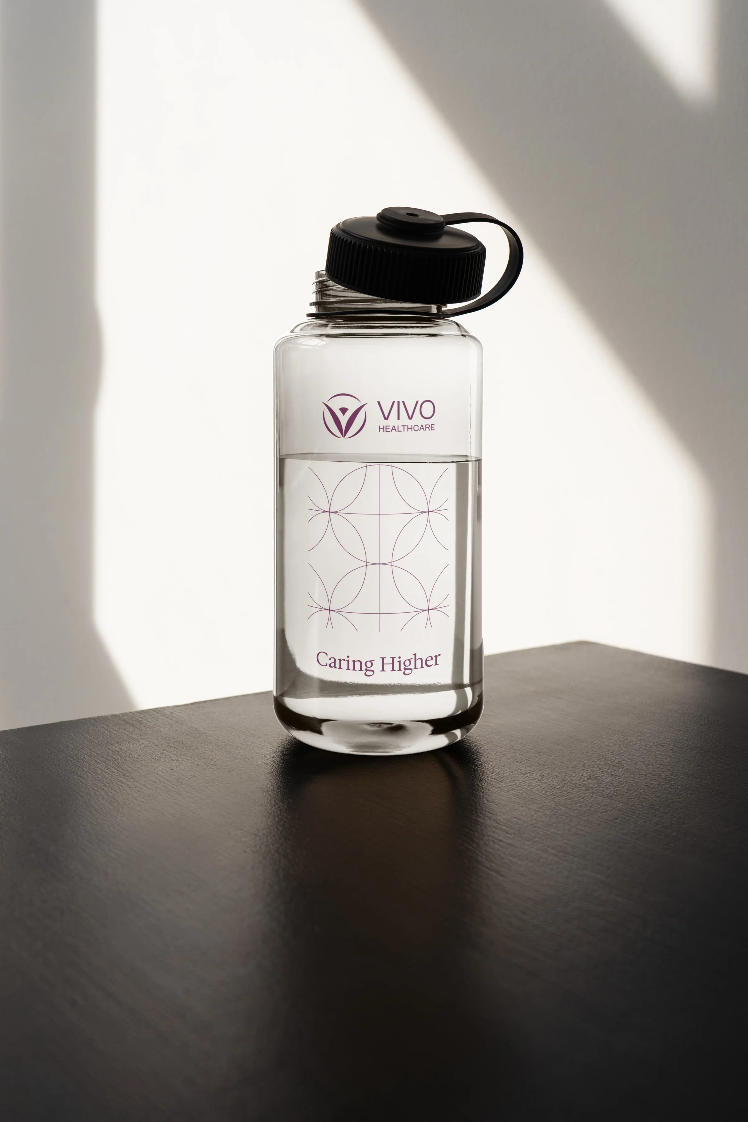 A clear Vivo Healthcare water bottle with the logo, the lattice pattern, and Caring Higher, on a dark table in sunlight