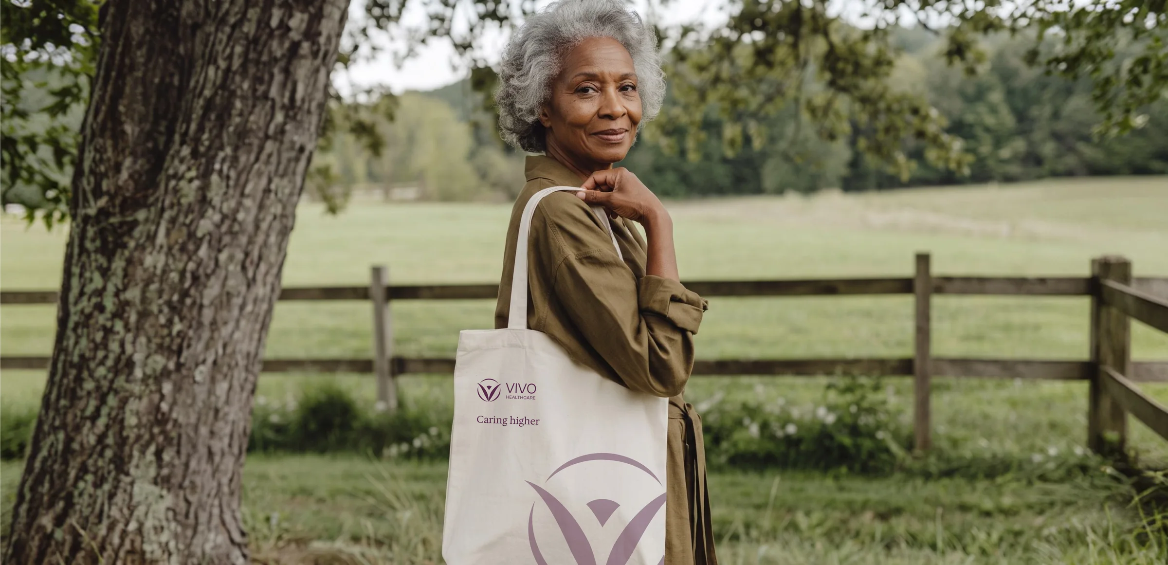 A woman leaning on a fence in a field, carrying a canvas Vivo Healthcare tote with the mark printed large in purple